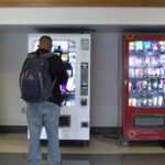 A male African-American student stands in front of the school supplies vending machine.