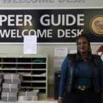 An African young woman stands in front of the Peer Guide Welcome Desk sign in Building 700.