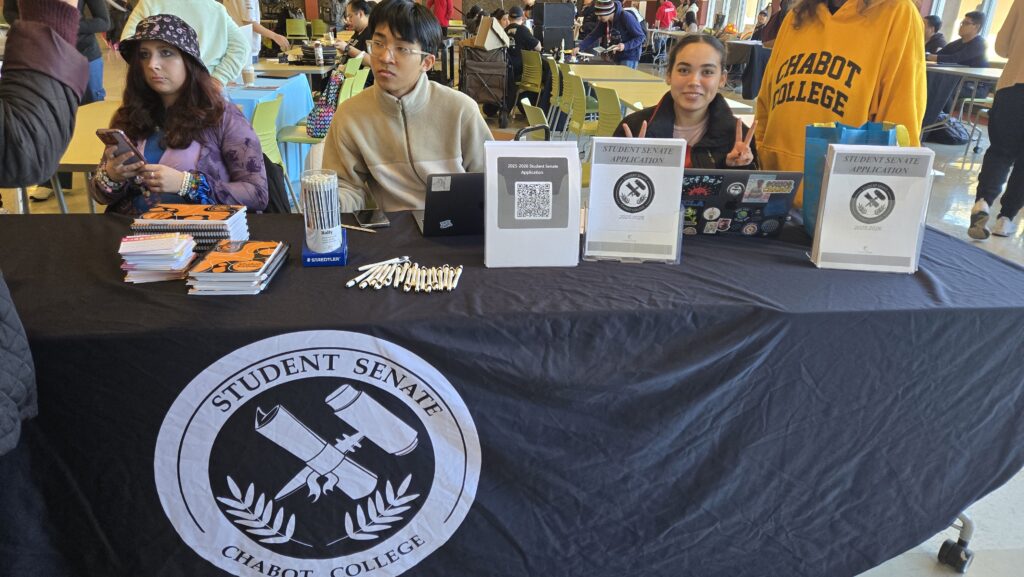 A group of students is sitting at a table representing the Student Senate of Chabot College in what appears to be a school event or fair. The table is covered with a black cloth featuring the Student Senate logo, which includes a gavel and scroll surrounded by laurel leaves. Various promotional materials, including notebooks, pens, and pamphlets, are neatly arranged on the table. There are also binders labeled “Student Senate Application” with QR codes for students to scan. One of the students is making a peace sign while smiling, and another is using a laptop. A person in a yellow “Chabot College” sweatshirt stands nearby, and several other students are visible in the background, engaged in various activities.