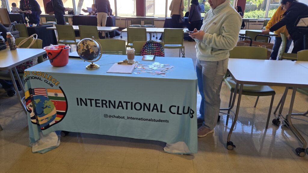 A table at an indoor event is covered with a blue tablecloth displaying “INTERNATIONAL CLUB” along with a logo featuring multiple country flags and a globe. The Instagram handle “@chabot_internationalstudents” is printed on the tablecloth. On the table, there is a small globe, a red bucket filled with candy, a clipboard with papers, a coffee cup, and an assortment of stickers. A person wearing a light-colored fleece jacket and jeans is standing next to the table, using their phone. In the background, other tables with people engaging in activities and conversations are visible, along with large windows allowing natural light into the room.