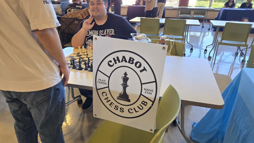 A table at an indoor event is set up for the Chabot Chess Club. A large sign with a black-and-white design features a chess piece and the words “CHABOT CHESS CLUB” along with the phrases “PLAY CHESS” and “HAVE FUN.” Behind the table, a smiling individual wearing a “Demon Slayer” T-shirt makes a peace sign while sitting in front of a chessboard with pieces set up for a game. Another person wearing a white shirt and jeans stands nearby, facing the table. In the background, other attendees and tables are visible, with chairs and event booths arranged in the room.