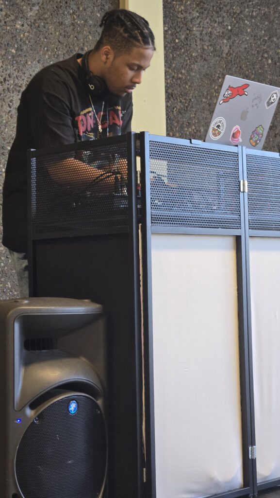 A DJ is performing at an event, standing behind a black DJ booth with a perforated metal design. He is wearing a black t-shirt with a graphic print, black headphones around his neck, and has braided hair. He is focused on his equipment, adjusting controls. A laptop with various stickers, including a red cartoon character and a skull, is placed on the booth. A large speaker is positioned in the foreground. The background features a textured wall, and the indoor setting suggests a social or community gathering.