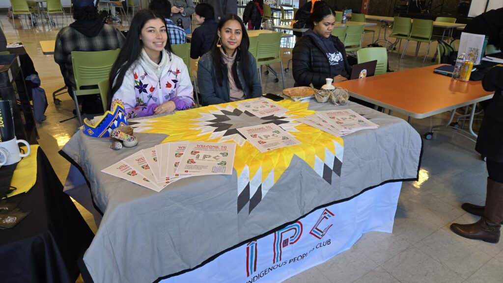 Two young women are seated at a table representing the Indigenous Peoples Club (IPC) at an event. The table is covered with a colorful quilt featuring a yellow, white, and black geometric design, with the club’s name embroidered at the bottom. Various informational flyers about the club are spread across the table, along with handcrafted items such as a beaded headdress and a small ceramic vase. One of the women is wearing a white jacket adorned with floral embroidery, while the other wears a dark jacket and a scarf. Both are smiling and engaging with event attendees. In the background, other people are seated at tables, including a third person working on a laptop at a nearby table.