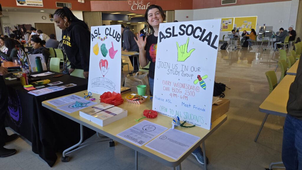 A smiling individual stands behind a table promoting the ASL (American Sign Language) Club at an event. They are making the “I love you” sign in ASL while standing next to two colorful, hand-drawn posters. One sign reads “ASL Social” and invites students to join a study group every Wednesday from 4 PM to 6 PM in a campus café. The table is covered with informational flyers, handmade paper flowers, and small decorations. Other attendees are engaging with different club tables in the background, and a student wearing a black hoodie with “Chabot College” printed on it is browsing a neighboring booth.