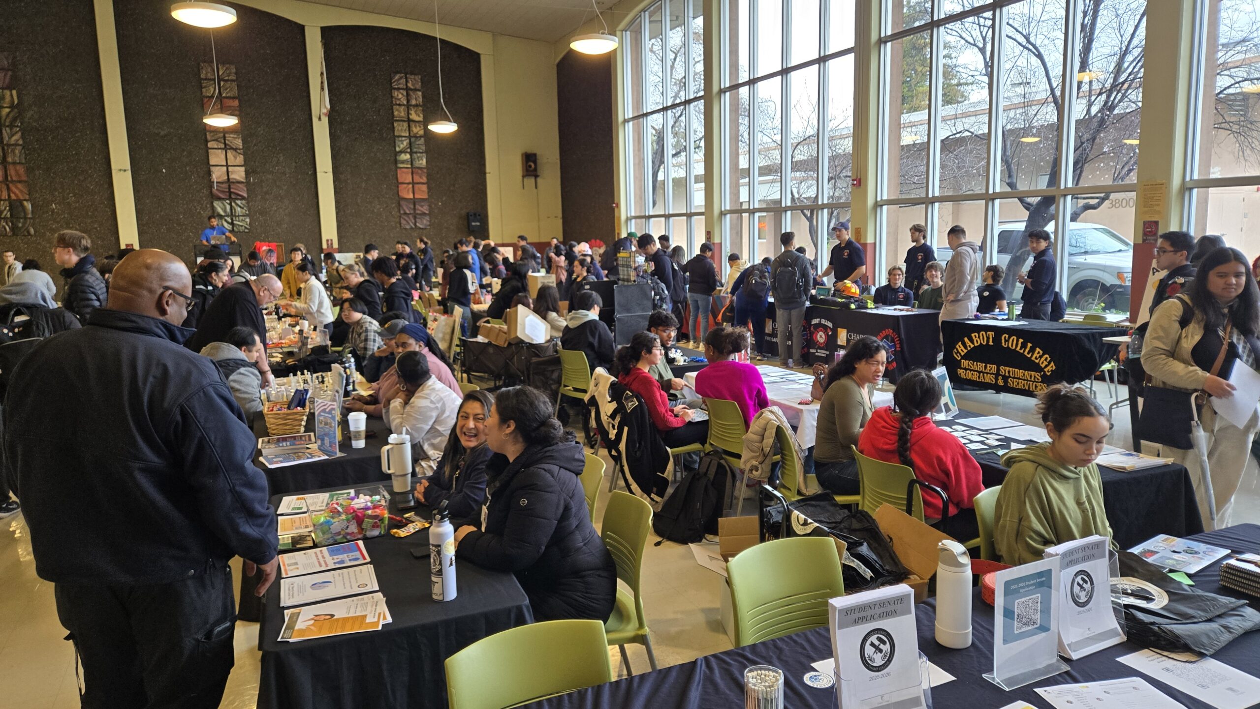 The image shows a room full of Chabot College students gathered around rows of tables draped in black tablecloths. Some students sit behind the tables and are giving information about various school clubs to the students who are standing in front of them.