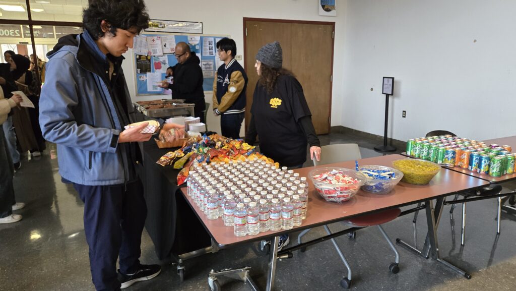 A student wearing a blue jacket is selecting food from a table filled with snacks, bottled water, and condiments. A person wearing a black “SSCC” shirt and a gray beanie stands behind the table, monitoring the setup. In the background, other attendees are seen near a food station where hot food is being served. Stacks of canned soda, chips, and various condiments are arranged on the tables, indicating a free food distribution event or a gathering with refreshments. A bulletin board with flyers and announcements is visible on the wall behind the setup.