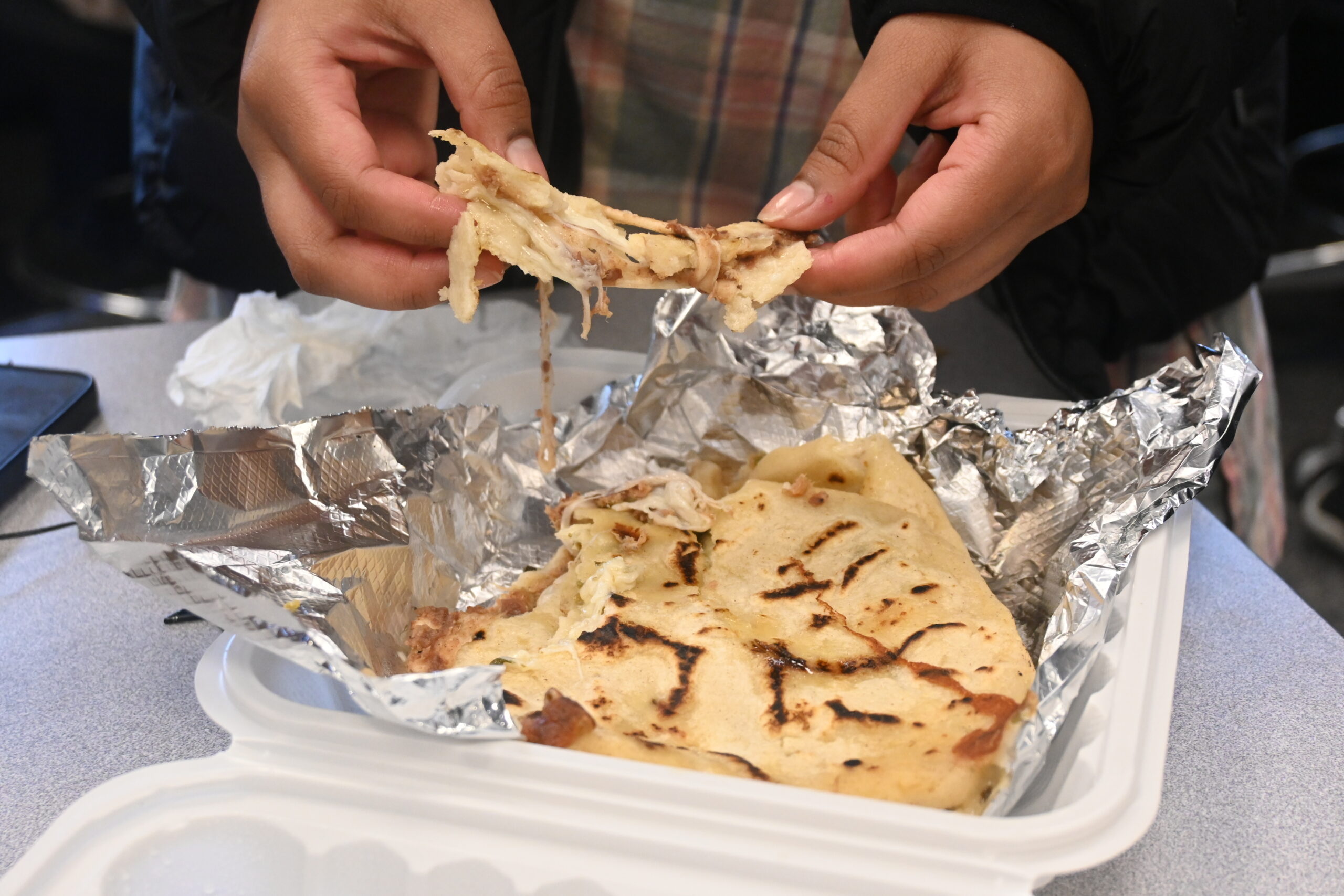 A pupusa being pulled apart with cheese falling out, aluminum in foil in a tray