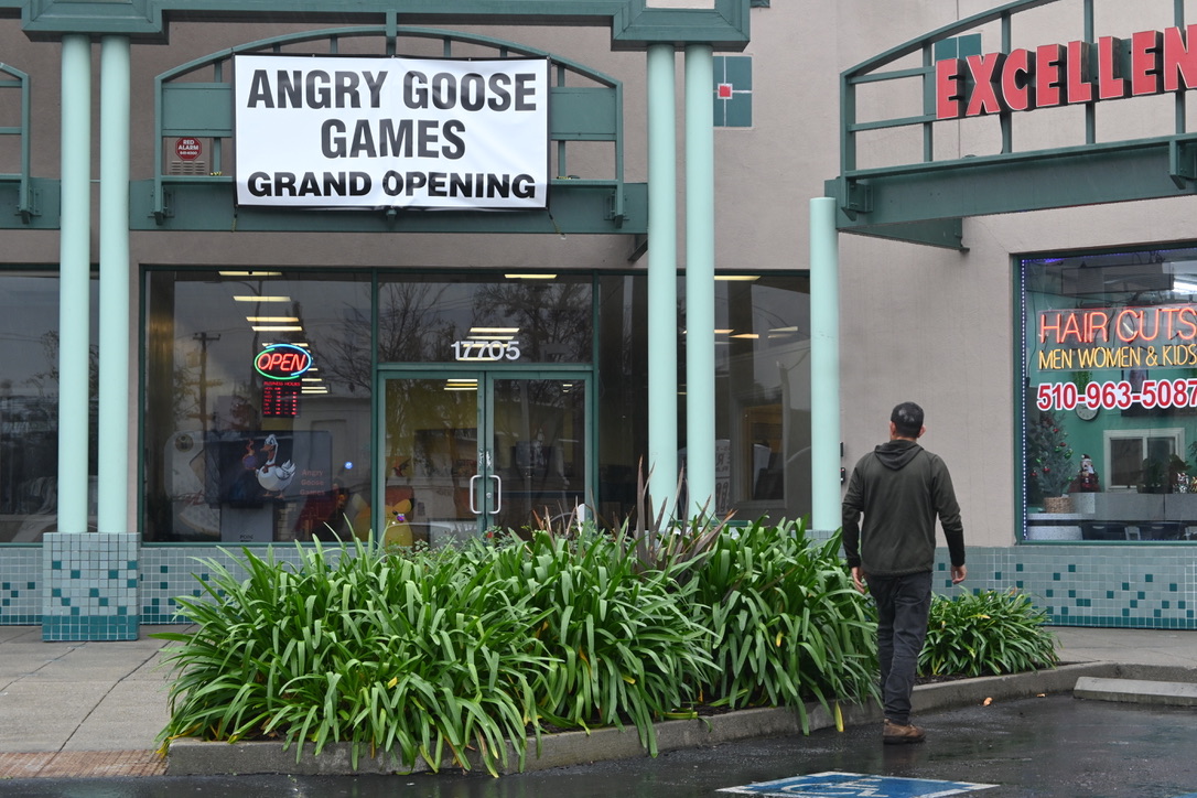 A man is walking up to a storefront in the rain. The storefront is green with large windows, and has a large white sign over the door with black text that reads "Angry Goose Games" in large font. Below that it says "Grand Opening" in a smaller font.